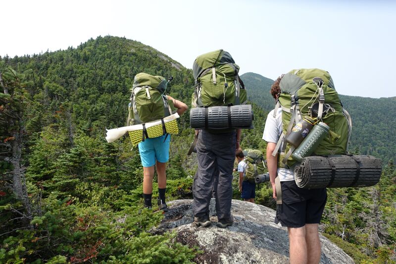The image shows three people hiking in a mountainous area. They are all wearing large backpacks, indicating they are likely on a multi-day trip. The surrounding landscape features lush green vegetation and a clear sky. The hikers appear to be enjoying the scenery as they make their way along the trail.